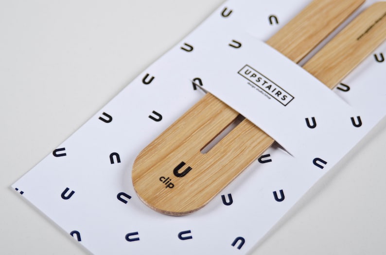May include: A wooden clip with the word "clip" and the letter "U" printed on it. The clip is on a white background with a pattern of the letter "U" repeated. The text "UPSTAIRS" is printed on a white label.