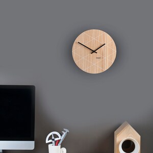Puede incluir: Un reloj de madera con un patr&oacute;n geom&eacute;trico blanco en una pared gris. El reloj tiene agujas negras y una esfera blanca. El reloj est&aacute; colgado sobre un escritorio con una computadora, una taza de l&aacute;pices y un objeto de madera con forma de casa.