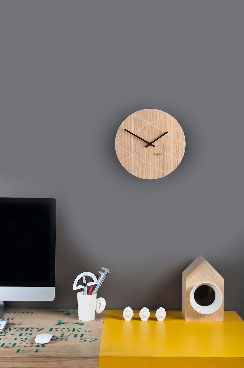 Puede incluir: Un reloj de madera con un patr&oacute;n geom&eacute;trico blanco en una pared gris. El reloj tiene agujas negras y una esfera blanca. El reloj est&aacute; colgado sobre un escritorio con una computadora, una taza de l&aacute;pices y un objeto de madera con forma de casa.