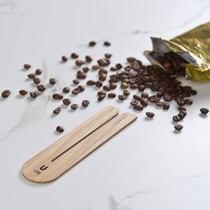 May include: A wooden coffee scoop with the text "U-clip" on it. The scoop is on a white marble surface with coffee beans scattered around it.