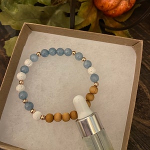 May include: A blue, white, and brown beaded bracelet with a small bottle of essential oil. The bracelet is in a brown gift box.