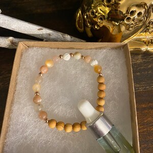 May include: A beaded bracelet with peach and white stones and wooden beads, presented in a small box with a glass dropper bottle. The bracelet features gold-colored accents. The background includes a gold-colored decorative object.