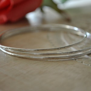 Puede incluir: Tres pulseras de plata apiladas una encima de la otra. Las pulseras son martilladas y tienen una superficie texturizada.