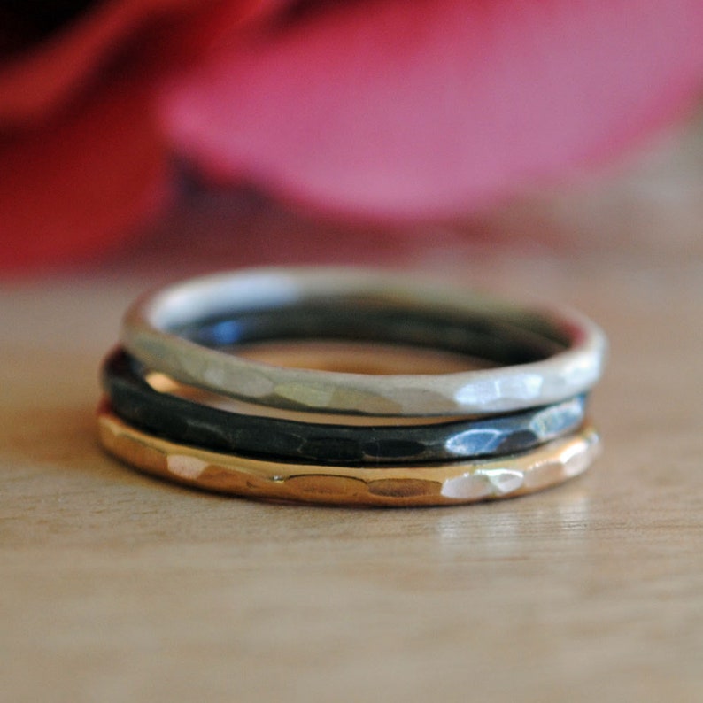 May include: Three thin hammered metal rings stacked on top of each other. The bottom ring is gold, the middle ring is black, and the top ring is silver.