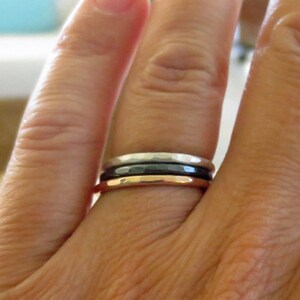 May include: A set of three stacking rings in silver, black, and rose gold. The rings are worn on a finger.