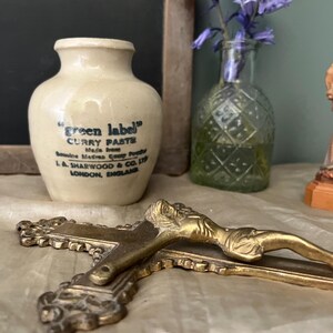 May include: A vintage ceramic jar with a white glaze and blue lettering that reads "green label" curry paste. The jar is sitting on a table with a small glass bottle of flowers and a wooden cross.