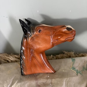 May include: A ceramic horse head sculpture with a glossy finish. The sculpture is mainly brown with black and white details, highlighting the horse's mane and facial features. The horse head is a decorative item.