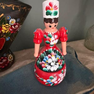 May include: A hand-painted wooden doll in a red dress and a white hat. The doll's dress is decorated with colourful floral designs in white, blue, and yellow. The doll has a painted face and arms.