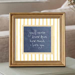 May include: A square gold-toned frame containing a print with a white and yellow vertical striped background. Centered is a dark blue denim-textured square with pinked edges featuring the white handwritten text: you'll never know dear how much i love you.
