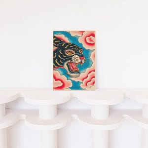 May include: A rectangular art print featuring a black panther head with yellow eyes and an open mouth showing sharp teeth. The panther is surrounded by stylized pink clouds against a bright blue background, all enclosed in a thin wooden frame.