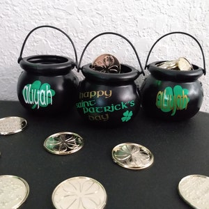 May include: Three black cauldron-shaped pots with arched handles, each containing coins. The pots are decorated with green shamrocks and the name "Aliyah" in white script. One pot has "Happy Saint Patrick's Day" in gold lettering.