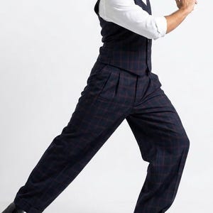 May include: A three-piece suit in a dark navy blue plaid pattern. The suit includes a vest, pleated trousers, and a white dress shirt. The trousers have a wide leg and are cuffed at the ankles. Black dress shoes complete the outfit.
