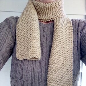 May include: A beige, hand-knitted scarf with a ribbed collar and a long, rectangular body. The scarf is made of a thick yarn, and the knit pattern is visible. The scarf is draped over a gray cable-knit sweater.