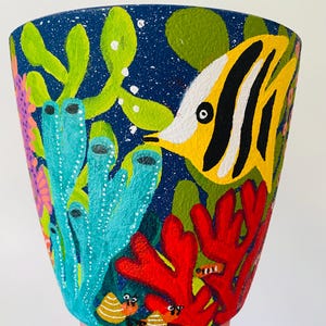 May include: Hand-painted ceramic pot depicting an underwater scene. It features a yellow and black striped fish, red coral, and various sea plants against a dark blue backdrop. The pot is held by a hand.