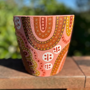 May include: A hand-painted terracotta flower pot with a textured surface. The pot features a pink base with a repeating pattern of brown, orange, and yellow abstract shapes. White, oval-shaped designs are also present. The pot is designed for indoor or outdoor use.