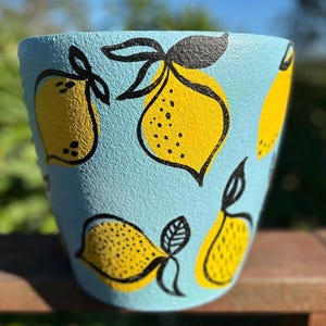 May include: A light blue ceramic plant pot with a textured surface. The pot is decorated with yellow lemons outlined in black. The lemons have black dots and leaves. Suitable for indoor or outdoor use.