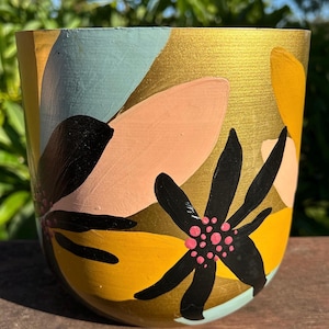 May include: A hand-painted ceramic planter with a gold metallic background and colourful floral designs. The flowers are black, yellow, peach, and light blue. The planter is a cylindrical shape.