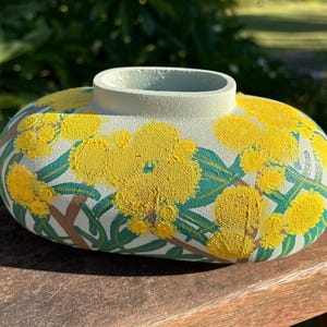 May include: A hand-painted ceramic vase with a pale green rim and body. The vase features yellow floral designs, green leaves, and brown stems. The oval-shaped vase is displayed on a wooden surface.
