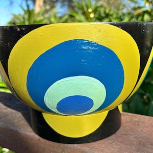 May include: A hand-painted wooden bowl with a black exterior and a vibrant interior design. The bowl features concentric circles in yellow, blue, and light blue. The bowl is set on a black base and is suitable for decorative use.