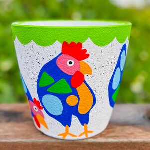 May include: A white ceramic plant pot with a textured finish, decorated with a hand-painted rooster design. The rooster is mainly blue with red, orange, and green details. The rim is green. Suitable for indoor or outdoor use.