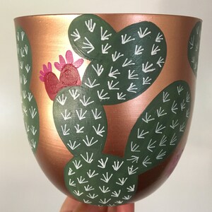 May include: Copper coloured ceramic planter with a hand-painted design of green cactus plants with white accents. The cactus plants are arranged in a pattern around the planter.