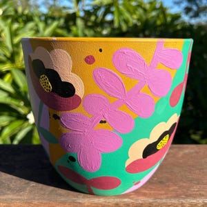 May include: A hand-painted ceramic plant pot with a vibrant floral design. The pot features a yellow background with pink, black, and cream-coloured flowers, and green and purple accents. The pot is round and sits on a wooden surface.