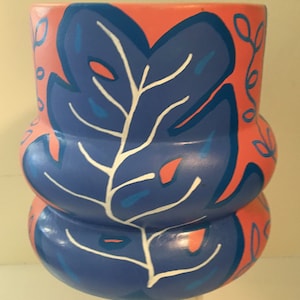 May include: A hand-painted ceramic vase with a blue and white leaf design on a coral background. The vase has a rounded, bulbous shape.