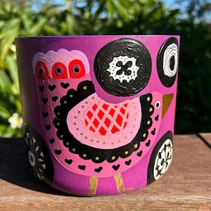 May include: A hand-painted, cylindrical planter in shades of purple, pink, black, and gold. The design features a stylized bird with a red and pink head, black accents, and gold legs. Decorative circles are painted around the bird.