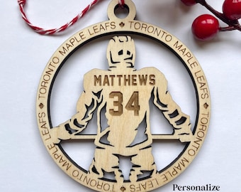 Puede incluir: Un adorno de jugador de hockey de madera con un diseño de Toronto Maple Leafs. El adorno está personalizado con el nombre "Matthews" y el número "34".