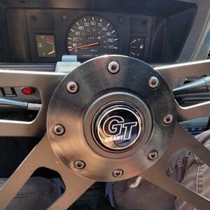 May include: Close-up of a black and silver Grant GT steering wheel in a vehicle. The steering wheel has a black center with the "GT Grant" logo and is surrounded by silver spokes. The dashboard and gauges are visible in the background.