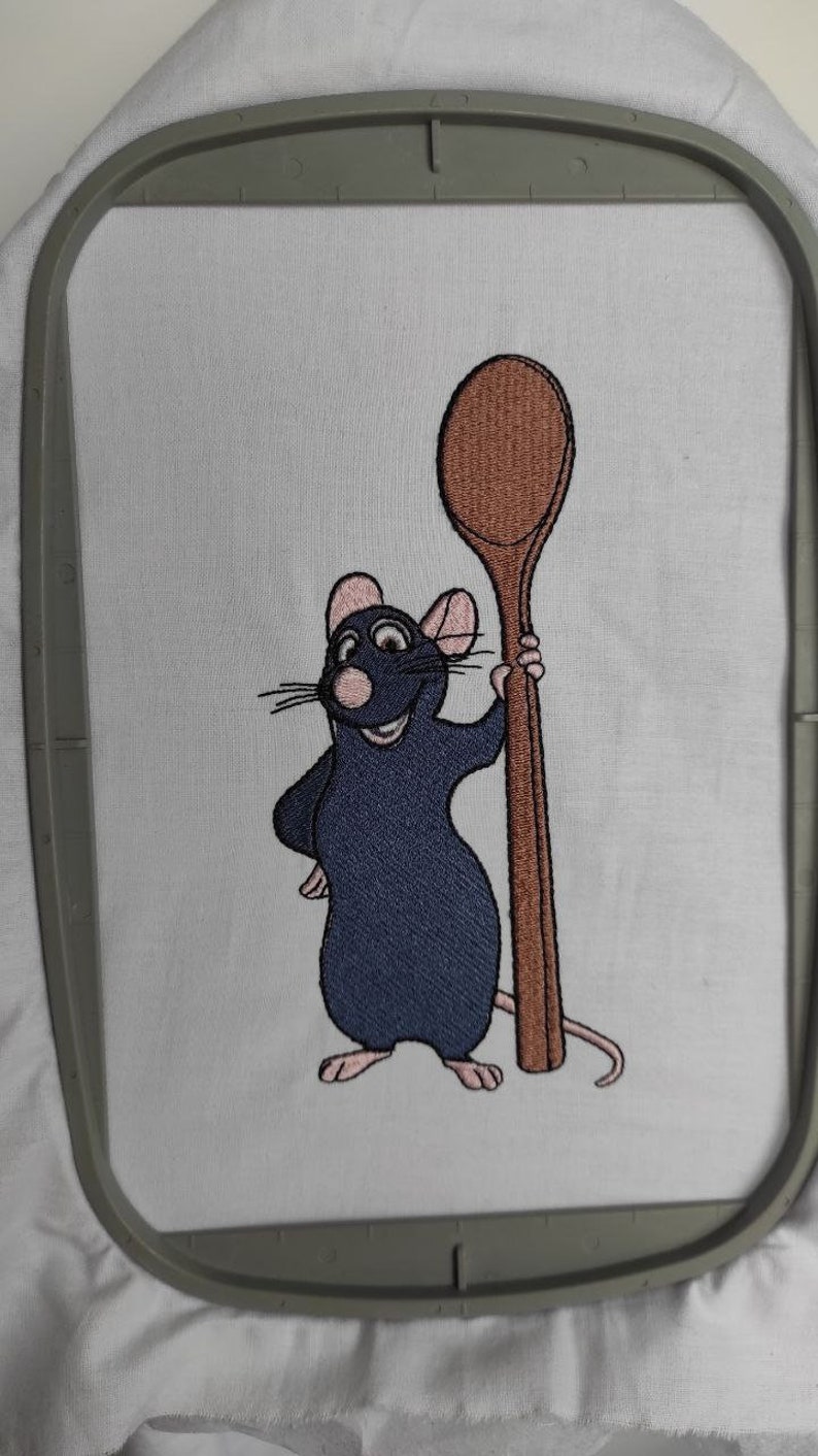 Puede incluir: Dise&ntilde;o bordado de una rata de dibujos animados sosteniendo una cuchara de madera. La rata es azul con orejas y nariz rosadas. La cuchara es marr&oacute;n.