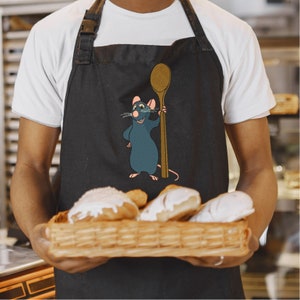 Puede incluir: Un delantal negro con una rata de dibujos animados sosteniendo una cuchara de madera. La rata es azul y tiene la nariz roja. El delantal lo lleva una persona que sostiene una cesta de pasteles.