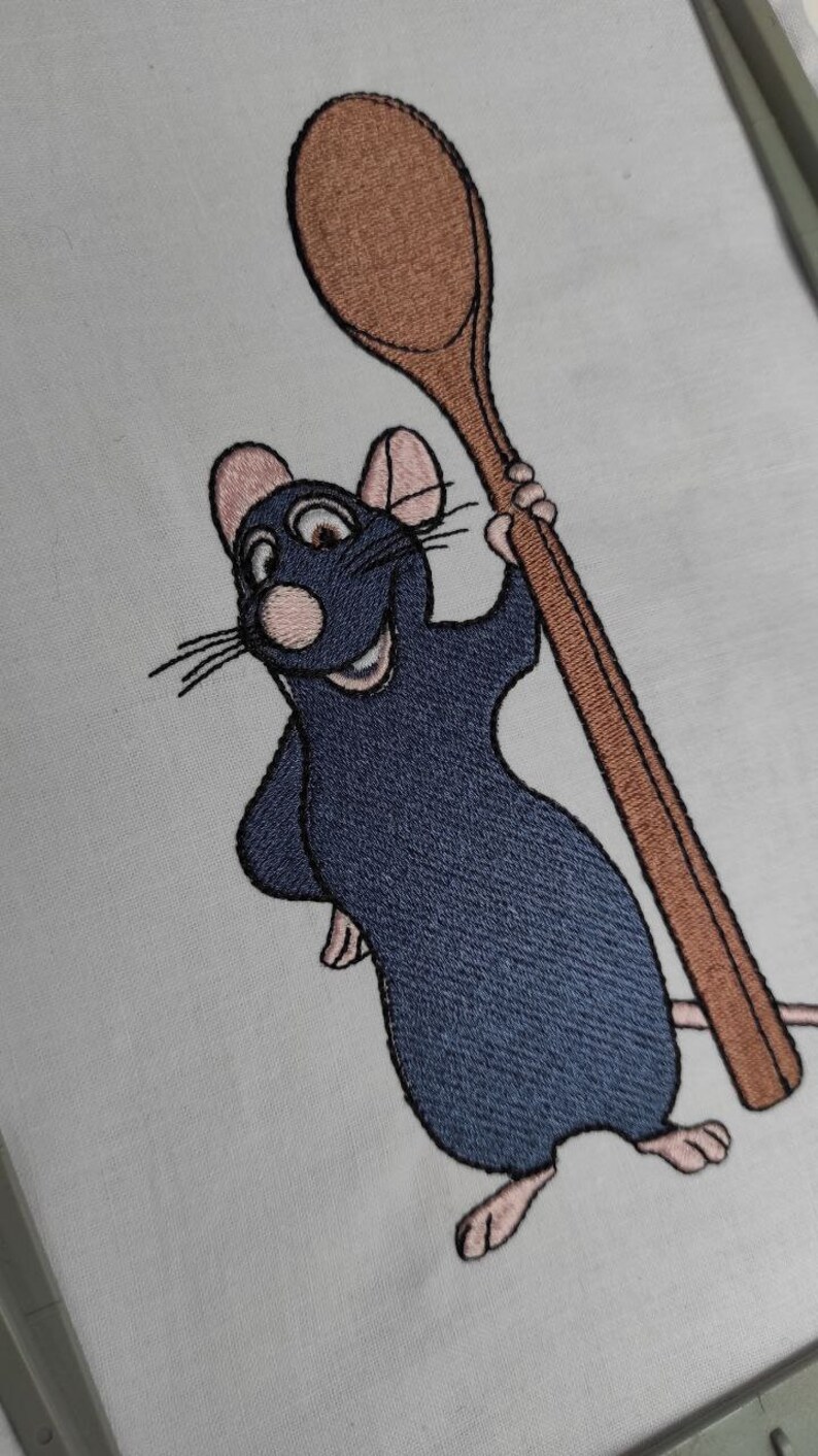 Puede incluir: Imagen bordada de una rata de dibujos animados sosteniendo una cuchara de madera. La rata es azul y tiene la nariz y las orejas rosas. La cuchara es marr&oacute;n.