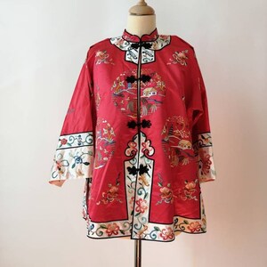 May include: A red silk jacket with intricate embroidery featuring floral patterns and a traditional Chinese landscape scene. The jacket has a mandarin collar and a front closure with black buttons.
