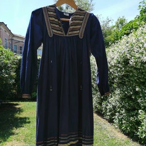 Puede incluir: Un vestido azul marino de manga larga con cuello en V y cuerpo fruncido. El vestido tiene un ribete de rayas marrones y beige alrededor del cuello y el dobladillo.