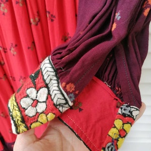 Puede incluir: Primer plano de un vestido rojo y burdeos con bordados florales. El vestido tiene una manga larga con un pu&ntilde;o fruncido y un cierre de bot&oacute;n. El bordado es en tonos de blanco, amarillo y negro.