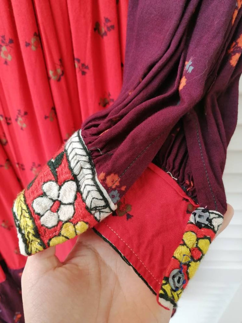Puede incluir: Primer plano de un vestido rojo y burdeos con bordados florales. El vestido tiene una manga larga con un pu&ntilde;o fruncido y un cierre de bot&oacute;n. El bordado es en tonos de blanco, amarillo y negro.