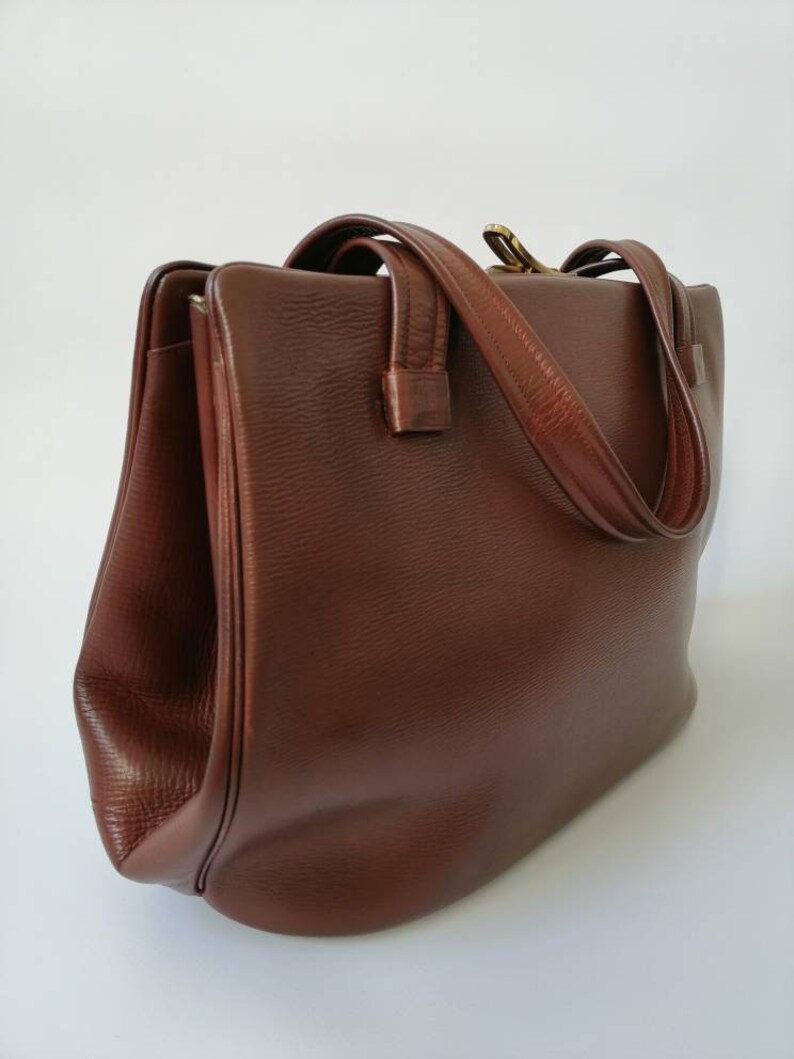 Puede incluir: Un bolso tote de cuero marr&oacute;n con dos asas superiores y un cierre de broche.