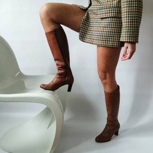 May include: A woman wearing a brown and white plaid blazer and brown knee-high boots is standing on a white chair.