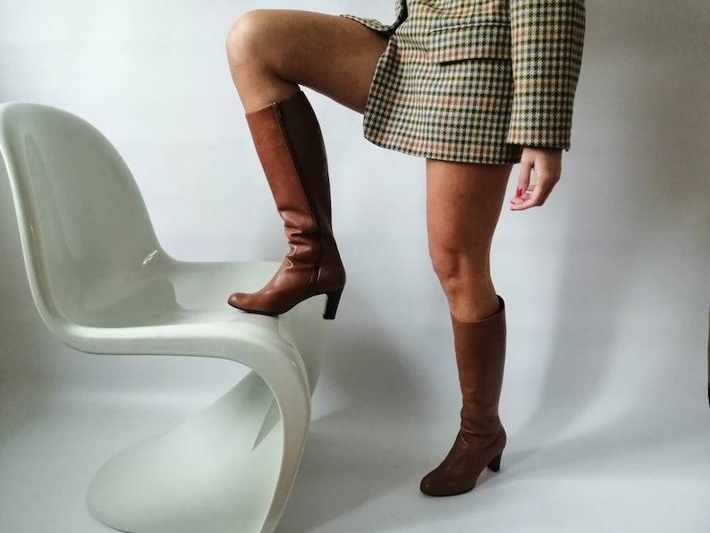 May include: A woman wearing a brown and white plaid blazer and brown knee-high boots is standing on a white chair.