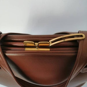 Puede incluir: Un bolso de mano de cuero marr&oacute;n con un cierre dorado y una sola correa. El bolso tiene una superficie texturizada y una forma ligeramente redondeada.
