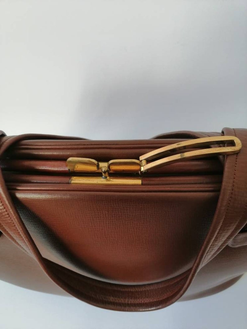 Puede incluir: Un bolso de mano de cuero marr&oacute;n con un cierre dorado y una sola correa. El bolso tiene una superficie texturizada y una forma ligeramente redondeada.