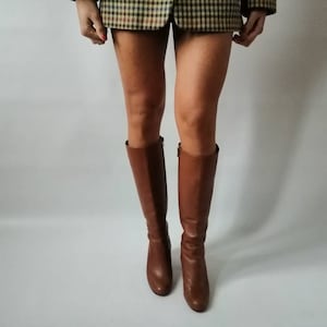 May include: Brown leather knee-high boots with a low heel. The boots are worn by a person wearing a plaid skirt.