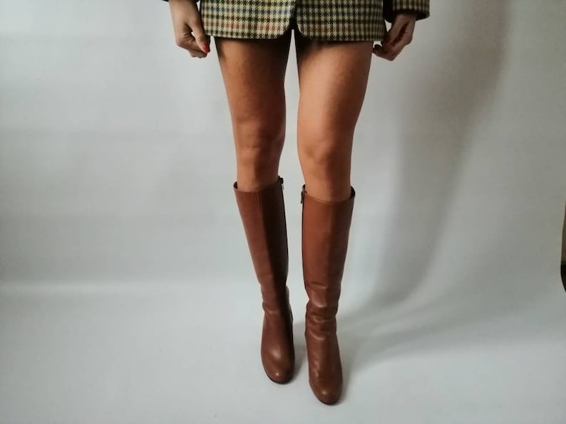 May include: Brown leather knee-high boots with a low heel. The boots are worn by a person wearing a plaid skirt.