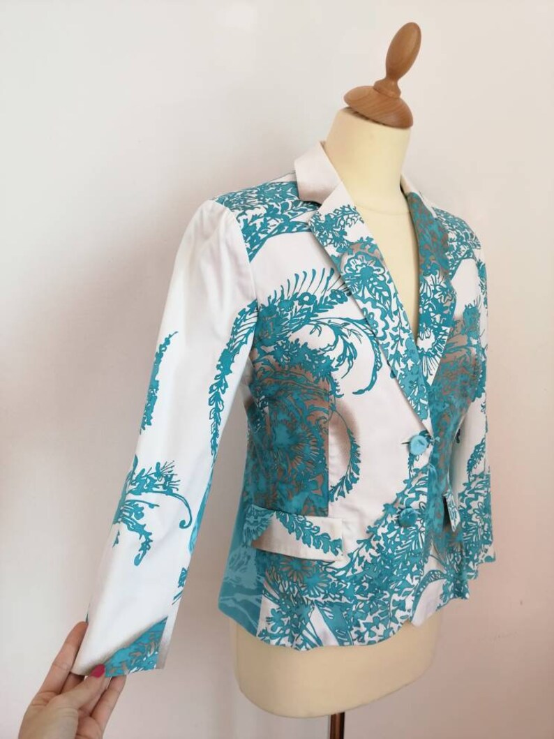 May include: A white blazer with a teal floral pattern. The blazer has a notched collar, long sleeves, and a single button closure.
