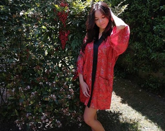 May include: A woman wearing a red and black silk robe with floral patterns. She is standing in a garden with green foliage and red berries.