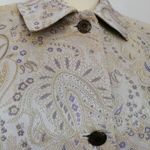 May include: A close-up of a light gray jacket with a paisley pattern in gold and purple. The jacket has a collar and buttons.