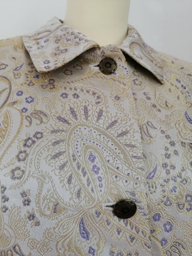 May include: A close-up of a light gray jacket with a paisley pattern in gold and purple. The jacket has a collar and buttons.