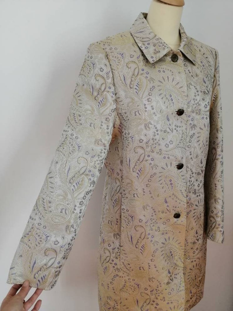 May include: A long-sleeved jacket with a paisley pattern in shades of gold, silver, and purple. The jacket has a collared neckline and a button closure.
