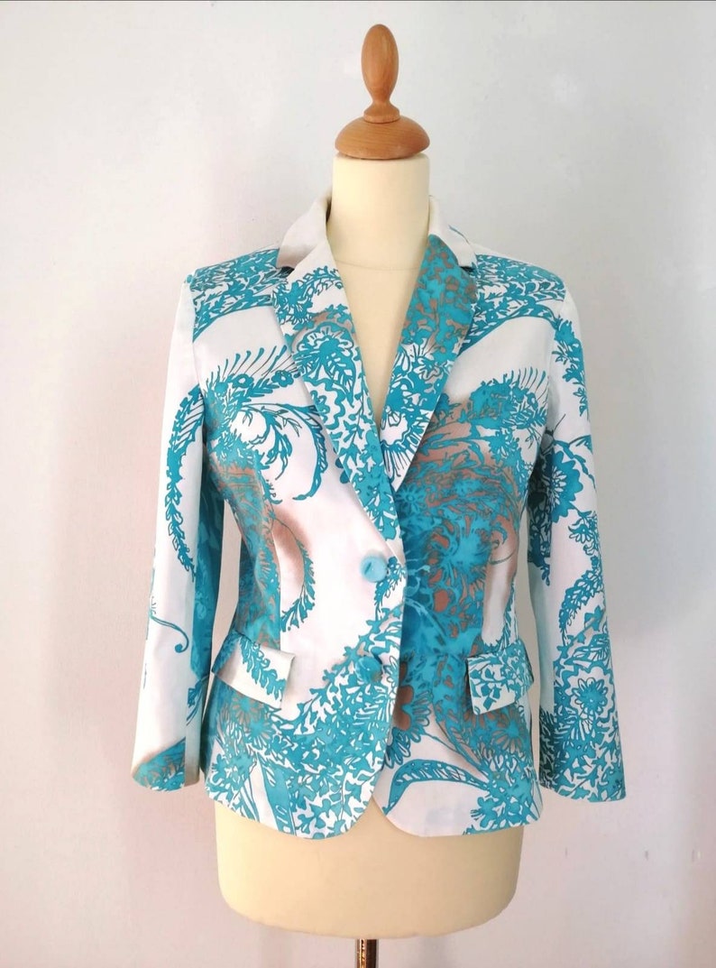 May include: A white blazer with a teal floral pattern. The blazer has a single button closure and a pocket on the left side.